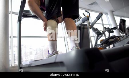 Mann mit Kniebandage leidet unter Schmerzen, steht auf Laufband, Rehabilitation Stockfoto