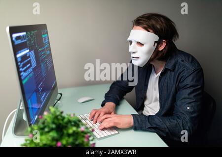 Anonymer Computerhacker in weißer Maske und Hoodie. Verdecktes dunkles Gesicht, das eine Geste zum Schweigen in der Secret Location im Underground Macht, Umgeben von Displays an Stockfoto