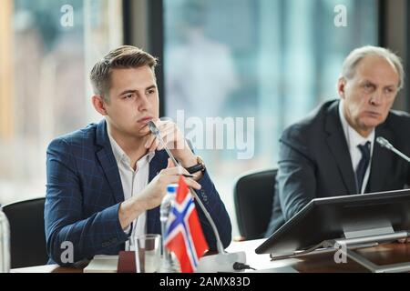 Junge ernsten politischen Führer in der Anzug sitzt mit seinem älteren Kollegen und hören bei Business Konferenz Stockfoto