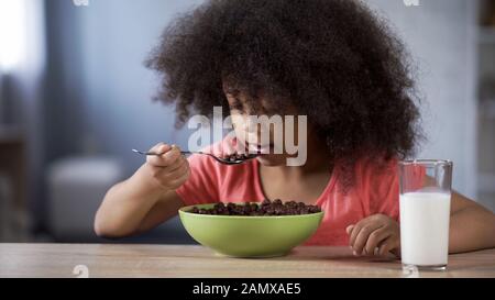 Entzückendes afrikanisches Mädchen, das Schokoladenkornflocken mit Appetit isst und Milch trinkt Stockfoto