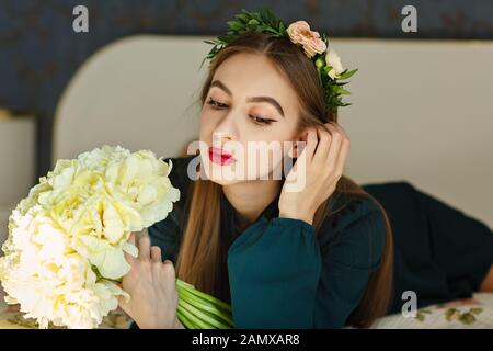 Portrait von Schönheit junge Mädchen in einer frischen Blume Kranz auf dem Kopf, auf dem Bett und hält einen großen Strauß gelber Blumen. Das Konzept der frisch Stockfoto