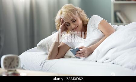 Lächelnde Frau im Bett liegend und E-Mail auf Smartphone, morgens Stockfoto