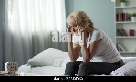 Seniorin sitzt auf dem Bett und leidet unter schrecklichen Kopfschmerzen, Gesundheitsproblem Stockfoto