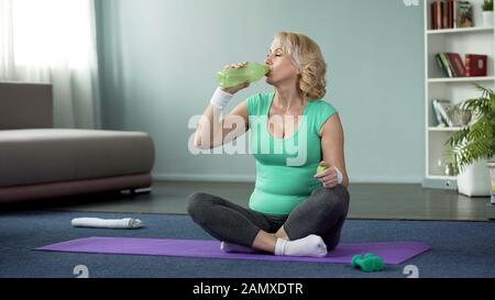 Athletische Seniorin sitzt nach dem Training auf dem Boden und trinkt Wasser, Gesundheit Stockfoto
