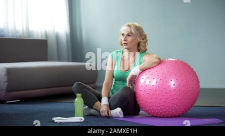 Müde, reife Frau, die nach dem Training auf einer Matte sitzt und sich am Fitball anlehnt Stockfoto