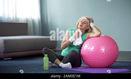 Ausgeschöpfte aktive Dame, die nach dem Heimtraining mit Fitnessball mit Handtuch wischt Stockfoto