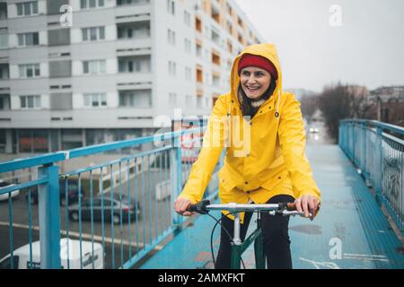 Lächelnde Frau Radfahren im Regen. Mädchen im gelben Regenmantel Radfahren Stockfoto