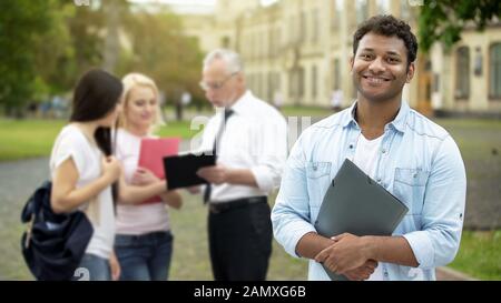Fröhlicher hispanischer Student steht in der Nähe des College und lächelt mit der Kamera, Bildung Stockfoto