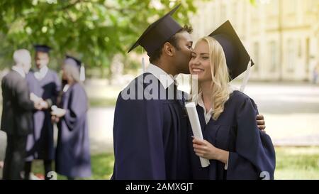 Absolventen der akademischen Regalien lächelnden, glücklichen Kerl, der Freundin auf Wange küsst Stockfoto