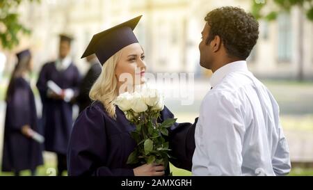 Weibliche Absolventin in akademischen Regalien mit Blumen sprechen mit Freund, Bildung Stockfoto
