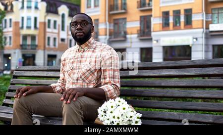 Nervöse junge Mann wartet auf Mädchen, sitzende Parkbank mit Blumen, blindes Datum Stockfoto