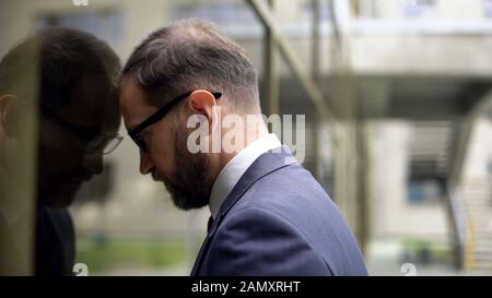 Erschöpfter Büroangestellter, der sich an die Glasbauwand lehnt, Depressionen Stockfoto