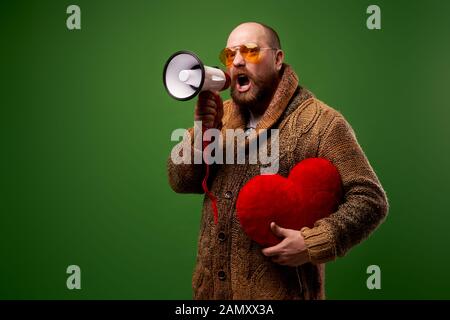 Mann in orangefarbenen Gläsern mit Spielzeugherz und Stierhorn in den Händen auf leerem grünem Hintergrund im Studio Stockfoto