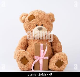 Große beige Teddybär mit Patches halten Geschenk in Safe in braunes Papier gewickelt, mit rosafarbenen Seidenband auf weißem Hintergrund gebunden, Happy Birthday und Valen Stockfoto