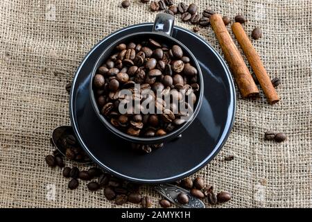 Schwarzer Kaffeebecher voll von organischem Kaffee Bohnen und Zimtstangen auf Leinen - Bild Stockfoto