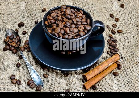 Schwarzer Kaffeebecher voll von organischem Kaffee Bohnen und Zimtstangen auf Leinen - Bild Stockfoto