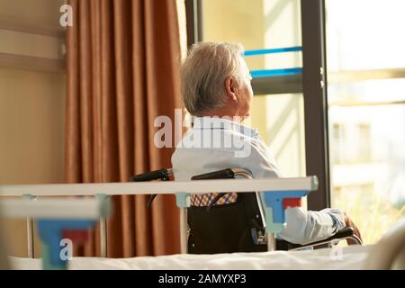 Rückansicht des älteren asiaten, der im Rollstuhl sitzt, in der Hausstation oder auf der Krankenstation, die aus dem Fenster blickt Stockfoto