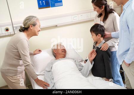 Glückliche asiatische Familie der drei Generationen, die mit dem hospitalisierten Großvater am Krankenbett spricht