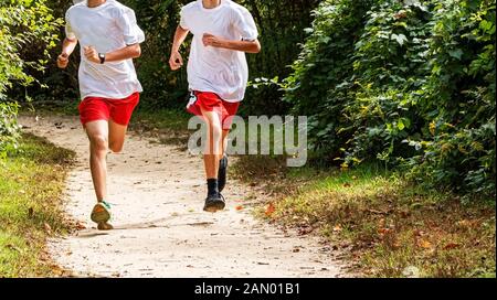 Zwei High School Querfeldeinläufer auf einem Training Laufen im Wald auf einem unbefestigten Weg tragen weiße Hemden und rote Shorts auf die Kamera läuft. Stockfoto