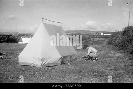 1950, historische, Camping, ein junger Mann, der in einem Feld tieing in den Boden und in den Stricken einer Leinwand, Zelt, in dem er gerade oben gesetzt hat, England, UK. Stockfoto