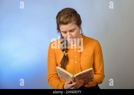 Ein Mädchen liest die Bibel beim Sitzen auf einem Stuhl auf einem blauen Hintergrund. Die Inschrift in Russisch - "Bibel" auf das Buch. Stockfoto