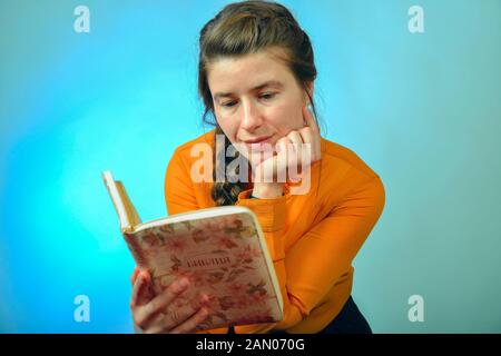 Ein Mädchen liest die Bibel beim Sitzen auf einem Stuhl auf einem blauen Hintergrund. Die Inschrift in Russisch - "Bibel" auf das Buch. Stockfoto