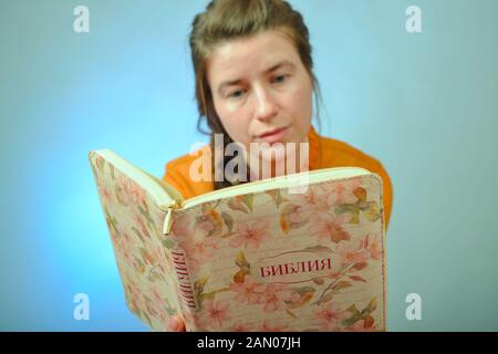 Ein Mädchen liest die Bibel beim Sitzen auf einem Stuhl auf einem blauen Hintergrund. Die Inschrift in Russisch - "Bibel" auf das Buch. Stockfoto