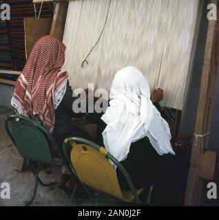 1960, historische, zwei arabische Männer sitzen auf Stühlen außerhalb, das Tragen der Kufiya, die traditionelle arabische Kopfbedeckung, mit der Hand an einem Holz gerahmt Webstuhl, ein Gerät, mit dem Tuch zu weben, Saudi-Arabien, Stockfoto