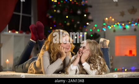 Junge Mutter und Tochter liegen unter dem Weihnachtsbaum und sehen sich an Stockfoto