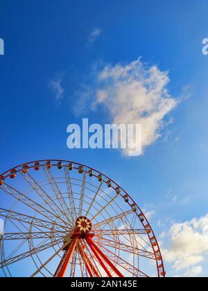 Teilweise sichtbares leuchtendes rotes Riesenrad mit roten, grünen und gelben Kabinen bei Sonnenuntergang. Keine Leute. Blauer Himmel mit weichen weißen Wolken. Vertikaler Hintergrund Wi Stockfoto