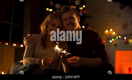 Süßes Paar mit bengalischen Lichtern, das in einem gemütlichen Zimmer in der Nähe eines dekorierten Weihnachtsbaums sitzt Stockfoto