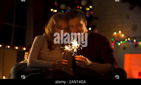 Liebevolles Paar mit bengalischen Lichtern, die in der Nähe des Weihnachtsbaums sitzen und sich in Neujahr wünschen Stockfoto