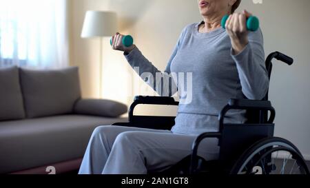 Seniorin im Rollstuhl, die Hanteln hebt, Übungen zu Hause macht, Genesung Stockfoto