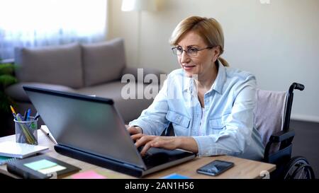 Erfolgreiche Seniorin im Rollstuhl arbeitet mit Laptop und chattet mit dem Kunden Stockfoto