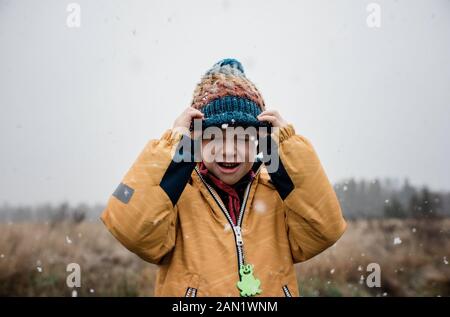 Junge zieht seinen Hut über sein Gesicht und lacht im Schnee Stockfoto