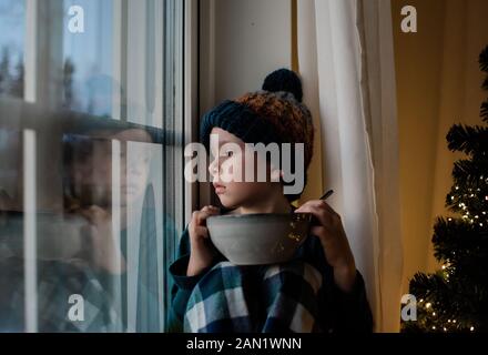 Der junge Junge saß auf einer Fensterbank mit einer Müslischale, die nach draußen schaute Stockfoto