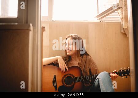 Fröhliche junge Frau, die Gitarre hält, während sie auf dem Balkonboden sitzt Stockfoto