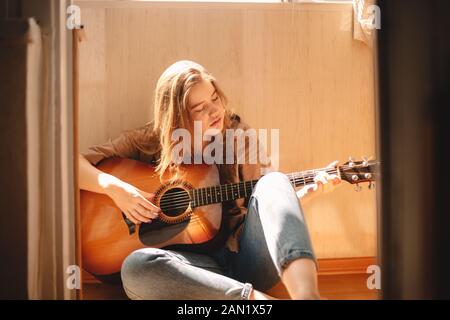 Junge Frau, die Gitarre spielt, während sie im Sommer auf dem Balkonboden sitzt Stockfoto