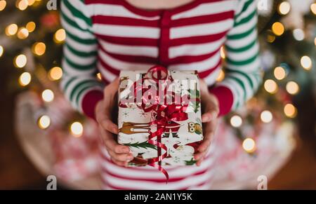 Kind im Pyjama hält Weihnachtsgeschenk vor Lichter auf Baum. Stockfoto