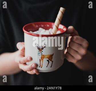 Nahaufnahme der Hände des Kindes, die heiße Schokolade in einem Rentierhausbecher halten. Stockfoto