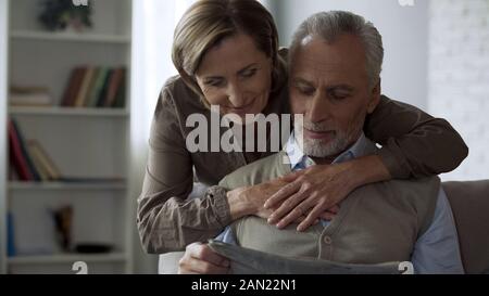 Alternde Dame ihr Mann umarmt, männlichen sitzen auf der Couch und liest Zeitung Liebe Stockfoto