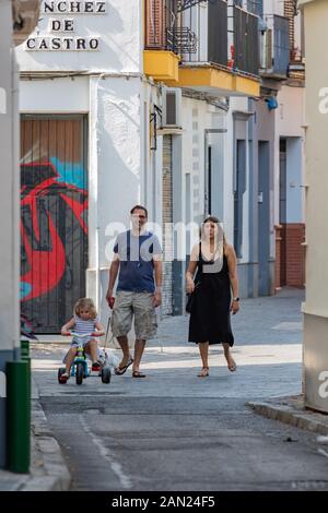 Eltern folgen geduldig einem kleinen Mädchen, das in einer ruhigen Straße in Sevilla auf ihrem Trike reitet Stockfoto