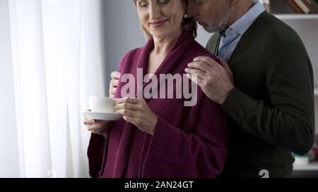 Im Alter von Lady Tee trinken stehen, Fenster, Mann umarmen und küssen geliebte Frau Stockfoto