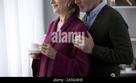 Im Alter von Paar in Fenster Frau Tasse Tee suchen, Mann sie hinter das Umarmen Stockfoto