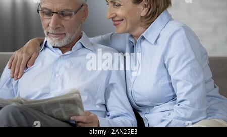 Rentner mann lesen Zeitung auf Sofa, Frau neben ihn umarmen, Liebe Stockfoto