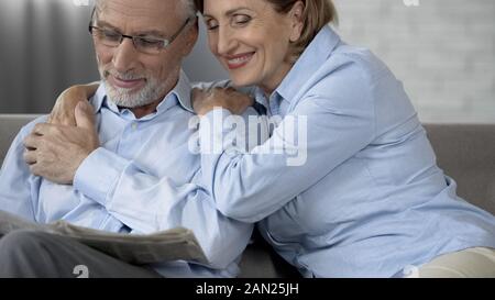 Alter Mann lesen Zeitung auf Couch, Frau saß neben ihm und umarmte und lächelnd Stockfoto