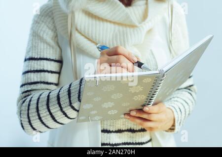 Nahaufnahme von stilvoller Frau in weiß gestreiftem Pullover, Schal und Ohrenklappenhut mit Schreibstift im Notizbuch vor hellblauem Hintergrund im Winter. Stockfoto