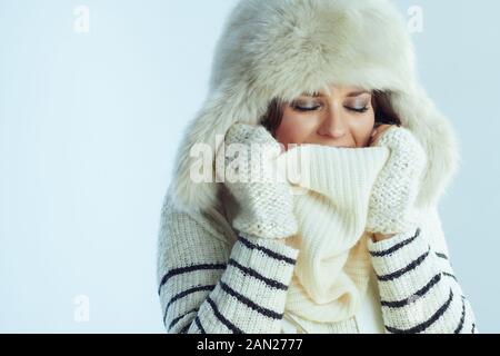 Gefrorene junge 40 Jahre alte Frau im weiß gestreiften Pullover und Ohrenklappen Hutbedeckung im Schal isoliert auf winterhellblauem Hintergrund. Stockfoto