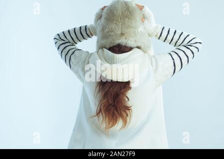 Von hinten gestresste Trendfrau im weiß gestreiften Pullover, Schal und Ohrenklappenhut auf winterhellblauem Grund. Stockfoto