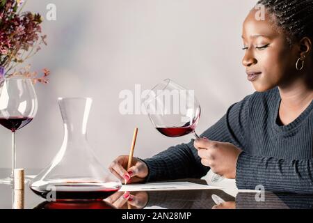 Close up Portrait von attraktiven afrikanische Frau mit Zöpfen Bewertung Rotwein auf dem Tisch. Mädchen Notizen neben Dekanter. Stockfoto